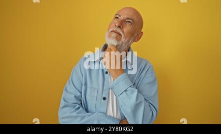 Ein Mann mittleren Alters mit grauen Haaren und Bart sieht nachdenklich vor einem isolierten gelben Hintergrund in einer blauen Jacke aus Stockfoto