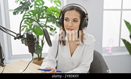 Eine junge hispanische Frau arbeitet in Innenräumen als Podcast-Moderatorin und spricht in einem hellen Studio mit Kopfhörern in ein Mikrofon. Stockfoto