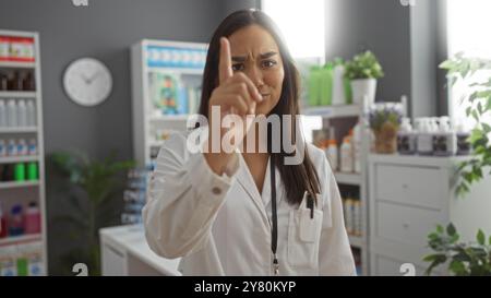 Junge brünette Frau in einer Apotheke zeigt mit dem Finger, während sie einen weißen Mantel trägt, mit Regalen von medizinischen Produkten im Hintergrund Stockfoto