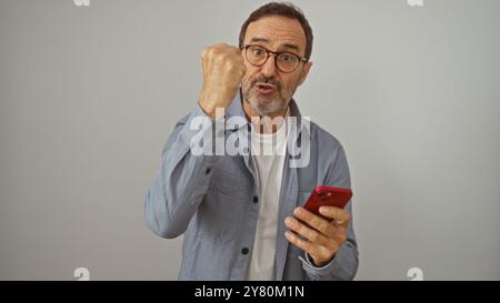 Hispanischer Mann feiert Erfolg mit erhobener Faust, während er ein Smartphone über isoliertem weißen Hintergrund hält. Stockfoto
