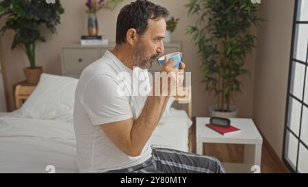 hispanischer Mann mittleren Alters sitzt auf einem Bett in einem gemütlichen Schlafzimmer, schlürft aus einem Becher in einem elegant dekorierten Innenraum mit Pflanzen und einem Nachttisch in einer Stunde Stockfoto