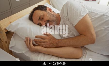 Gutaussehender hispanischer Mann mittleren Alters, der warm lächelt, während er im Bett lag und sich bequem in seinem Schlafzimmer ausruhte Stockfoto