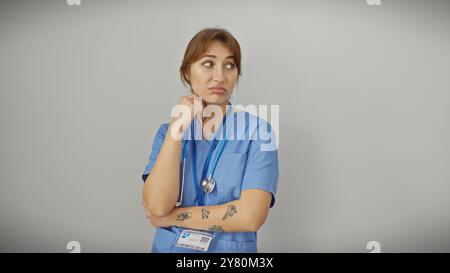 Nachdenkliche junge kaukasische Frau in blauen Schutteln mit Stethoskop, Tattoos und Abzeichen, isoliert auf weißem Hintergrund. Stockfoto