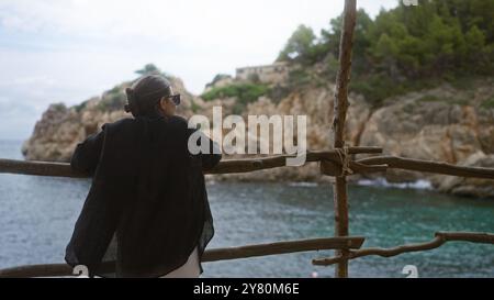 Junge Frau, die einen malerischen Blick auf das Meer in cala deia auf mallorca, spanien genießt, draußen am Holzzaun steht und auf die felsige Küste blickt Stockfoto