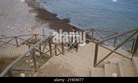 Junge Frau, die eine Holztreppe am felsigen Strand am Port de valldemossa in mallorca, spanien, hinuntergeht, mit dem klaren blauen Meer im Hintergrund Stockfoto