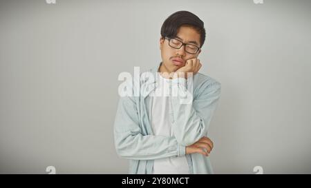 Ein junger asiatischer Mann mit Brille steht gelangweilt vor einem weißen, isolierten Hintergrund und zeigt eine lässige und moderne Atmosphäre. Stockfoto