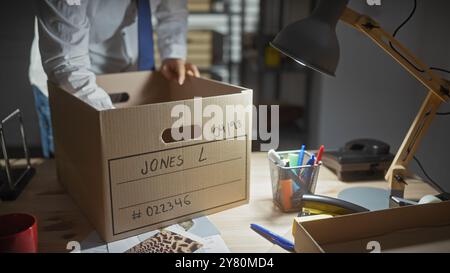 Ein Mann in einem Büro organisiert eine Pappschachtel mit Beweismitteln auf einem überfüllten Schreibtisch unter Lampenlicht. Stockfoto