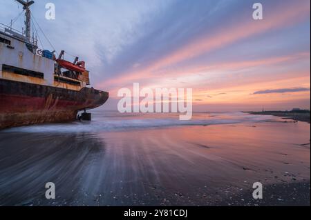 Ein verwittertes Schiff ist vor einem feurigen Sonnenuntergangshimmel auf dem goldenen Sand des Xigushui Beach geschildert. Der Kontrast zwischen dem lebendigen Himmel und dem dunklen Sch Stockfoto