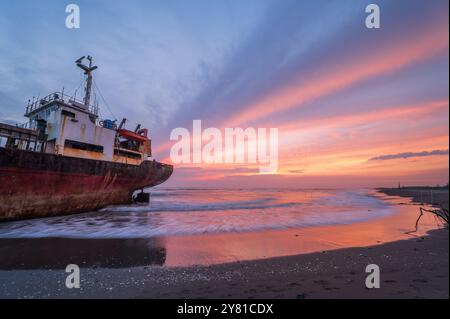 Ein verwittertes Schiff ist vor einem feurigen Sonnenuntergangshimmel auf dem goldenen Sand des Xigushui Beach geschildert. Der Kontrast zwischen dem lebendigen Himmel und dem dunklen Sch Stockfoto