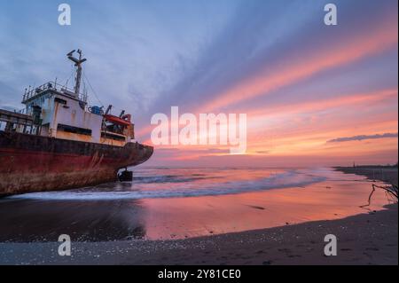 Ein verwittertes Schiff ist vor einem feurigen Sonnenuntergangshimmel auf dem goldenen Sand des Xigushui Beach geschildert. Der Kontrast zwischen dem lebendigen Himmel und dem dunklen Sch Stockfoto