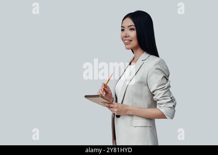 Alles organisieren. Attraktive junge asiatische Frau, die wegschaut und lächelt, während sie vor grauem Hintergrund steht Stockfoto