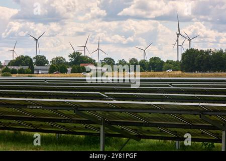 Ein großer Solarpark mit Reihen von Photovoltaik-Panels, neben einer Autobahn, am Rande einer Stadt, mit Bäumen, Windturbinen und bewölktem Himmel. Re Stockfoto