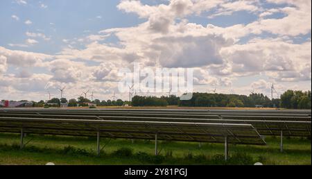 Ein großer Solarpark mit Reihen von Photovoltaik-Panels, neben einer Autobahn, am Rande einer Stadt, mit Bäumen, Windturbinen und bewölktem Himmel. Re Stockfoto