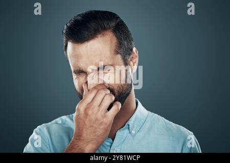 Allergie. Hübscher junger Mann, der die Nase mit der Hand bedeckt, während er vor grauem Hintergrund steht Stockfoto