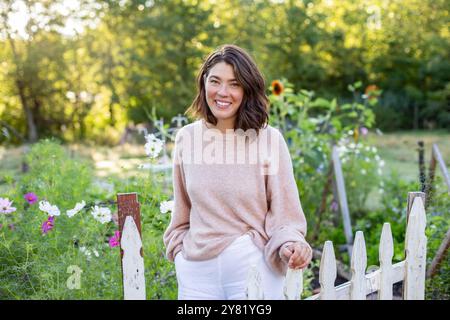 Frau lächelt in einem Garten mit einem Holzzaun und Blumen im Hintergrund. Stockfoto