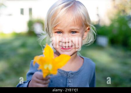 Lächelndes Kleinkind mit einer gelben Blume im Freien und einem Haus im Hintergrund. Stockfoto