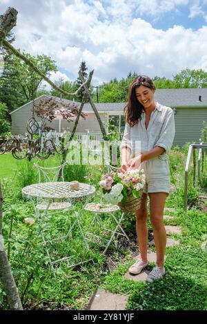 Lächelnde Frau, die Blumen in einem Korb neben einem weißen Metalltisch in einem sonnigen Garten arrangiert. Stockfoto