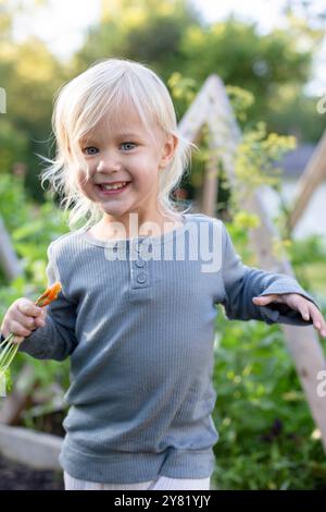 Lächelndes, blondes Kleinkind mit Karotten in sonniger Gartenumgebung. Stockfoto