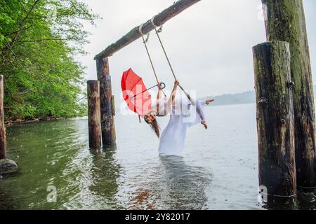 Frau, die an einem Seil über einem See schwingt und an einem nebeligen Tag einen roten Schirm hält. Stockfoto