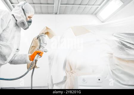 Automechaniker, der während der Reparaturarbeiten ein weißes Auto mit einer Spritzpistole in einer Lackkammer lackiert. Stockfoto