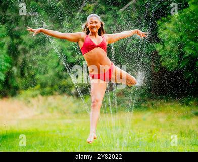 Lächelnde Frau in rotem Badeanzug, die an einem sonnigen Tag einen Sprenger genießt Stockfoto