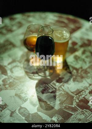 Ein Glas Rotwein und ein Pint Bier auf einem Tisch mit einer Collage aus schwarz-weißen Bildern unter einer Glasplatte. Stockfoto