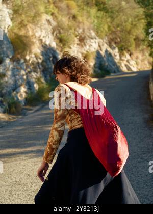 Eine Frau geht auf einer malerischen Straße mit einer roten Tasche, gekleidet in einem einzigartigen gemusterten Oberteil und einem schwarzen Rock. Stockfoto