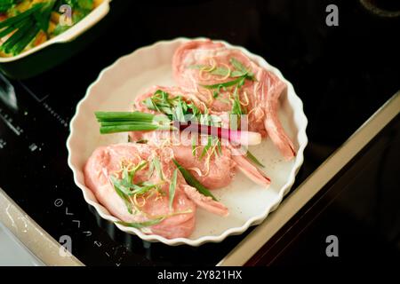 Frische, rohe Lammkoteletts, garniert mit Frühlingszwiebeln und Kräutern auf einem weißen Teller, fertig zum Garen. Stockfoto