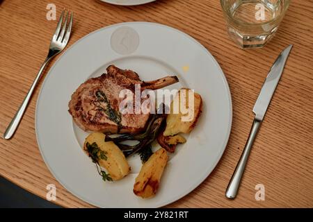 Gegrilltes Schweinekotelett, serviert mit gebratenen Kartoffeln auf einem weißen Teller, begleitet von einer Gabel, einem Messer und einem Glas Wasser auf einem Holztisch. Stockfoto