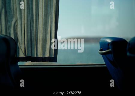 Innenansicht eines Busses mit einem Vorhang, der neben einem Fenster mit Blick auf das Meer und einen fernen Hügel unter klarem Himmel flattert. Stockfoto