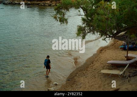 Eine einsame Figur spaziert an einem ruhigen Sandstrand neben ruhigem Wasser mit einem sanft überhängenden Baum und einer leeren Sonnenliege in der Nähe. Stockfoto