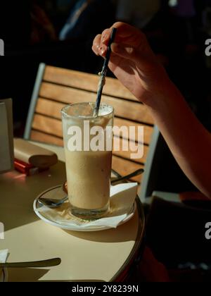 Rühren Sie ein hohes Glas cremigen Eiskaffee von Hand auf einem sonnigen Kaffeetisch. Stockfoto