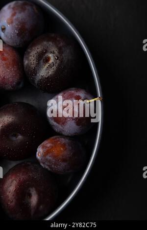 Saftige reife frische Pflaume mit Wassertropfen in grauer Platte auf schwarzem Hintergrund. Obst der Saison. Selektiver Fokus, Nahaufnahme. Stockfoto