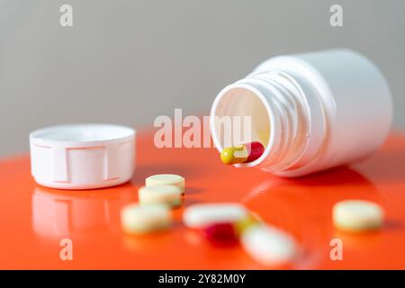 Eine weiße Pillenflasche mit abgezogenem Verschluss, die auf der Seite liegt und verschiedene Pillen und Kapseln auf einer orangefarbenen Oberfläche auslaufen. Stockfoto