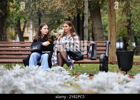 Zwei glückliche Frauen, die emotional auf einer Bank im Stadtpark im Herbst sprechen, Mädchen mit überraschtem Gesichtsausdruck Stockfoto