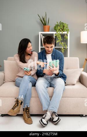 Entspanntes, junges Paar mit Smartphone und Tablet, das zu Hause auf einer Couch im Wohnzimmer sitzt. Diverse Paare, die lächeln und Spaß haben Stockfoto