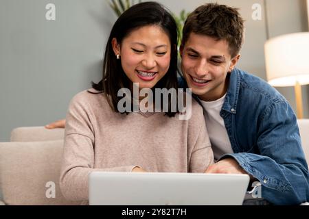 Glückliches junges, entspanntes, vielfältiges Paar, das auf dem Sofa sitzt und einen Laptop benutzt. Liebevoll aufgeregte Frau und Mann mit Computer im Wohnzimmer. Stockfoto