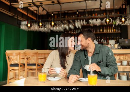 Ein Paar teilt sich einen freudigen Moment in einem stilvollen Café, schlürft Kaffee und genießt hellen Orangensaft, während es gemeinsam lacht, umgeben von einem einladenden Restaurant Stockfoto