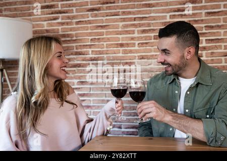 Lachender Mann und Frau, die zu Hause mit einer Brille trinken. Fröhliches junges Erwachsenenpaar, das mit Wein am Tisch toast. Stockfoto
