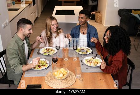 Lächelnde Leute, die Hände halten, bevor sie in einer modernen Wohnung essen. Verschiedene Gruppen glücklicher Freunde beten gemeinsam zu Hause. Stockfoto