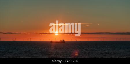 Silhouette eines Transportschiffes und Windparks in der Nordsee bei Sonnenuntergang. Silhouette eines Transportschiffes und Windparks in der Nordsee bei Sonnenuntergang. Ein Wolkenband in der Nähe der Sonne. Strand Scheveningen, Niederlande. niederlande den haag B97A7472 Stockfoto