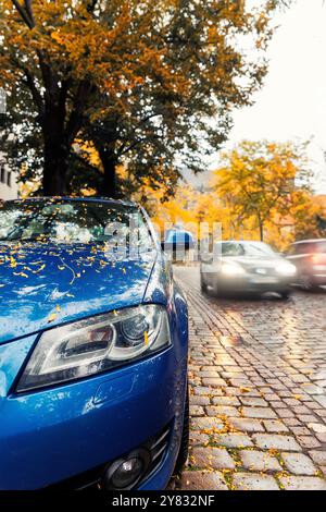 Moody, malerischer Blick auf die Frontscheinwerfer modernes weißes Auto Windschutzscheibe bedeckt goldene gelbe gefallene Blätter geparkt nasse Stadt Straße Herbst regnerischer Tag. Bewölkt Stockfoto