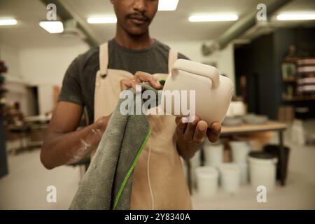 Nahaufnahme eines jungen afrikanischen Töpfers mit einem Tuch, um einen Tonbecher zu reinigen, während er in einem großen Keramikfabrikstudio arbeitete Stockfoto