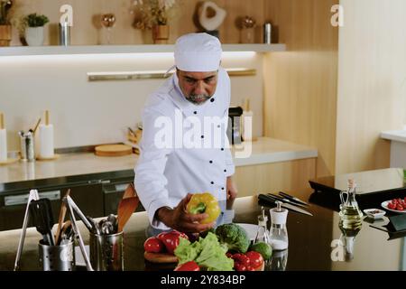 Indischer reifer Küchenchef in Uniform gekleidet mit gelbem Paprika in moderner professioneller Küche Stockfoto
