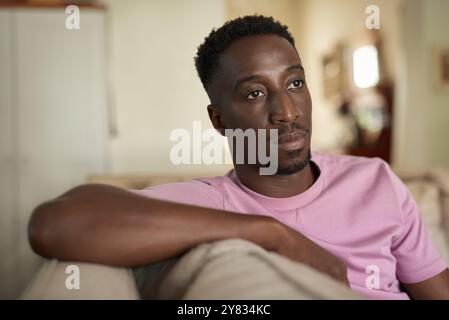 Junger afrikanischer Mann, der tief in Gedanken schaut, während er allein auf seinem Wohnzimmer-Sofa sitzt Stockfoto