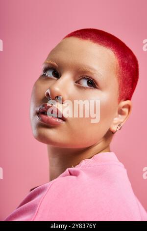 Eine kühne junge Frau zeigt ihren einzigartigen Stil mit kurzen rosa Haaren und einem Nasenstechen vor einem hellen Hintergrund. Stockfoto