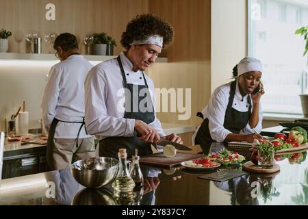 Professionelles Koch-Team in weißer Uniform mit schwarzen Schürzen, die Zutaten für Caprese-Vorspeise vorbereiten, afroamerikanische Köchin, die mit dem Kunden telefoniert und Details aufschreibt Stockfoto