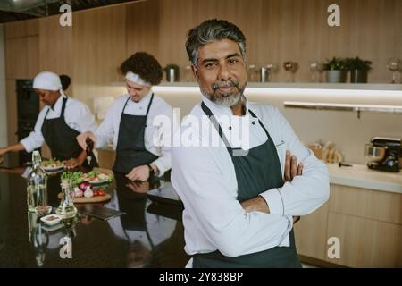 Porträt des Reifen indischen Küchenchefs in weißer Uniform, der leicht lächelt, während er in der Küche des eigenen Restaurants für ein Foto posiert Stockfoto