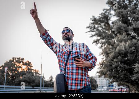 Porträt eines gutaussehenden Geschäftsmannes mit Sonnenbrille und kariertem Hemd, der auf der Straße spaziert und winkt, um das Taxi zu schnappen. Reifer, lässig gekleideter Mann mit Stockfoto