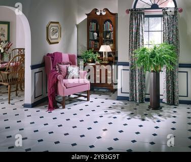 Helles traditionelles Wohnzimmer mit rosafarbenem Sessel, floralen Vorhängen, gefliestem Boden, Holzschrank, üppigem Farn durch sonnenbeleuchtetes Fenster in gemütlicher klassischer Einrichtung Stockfoto
